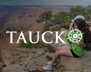 Two people embrace as they sit on a cliff looking over a grand valley, with the Tauck logo superimposed over them.
