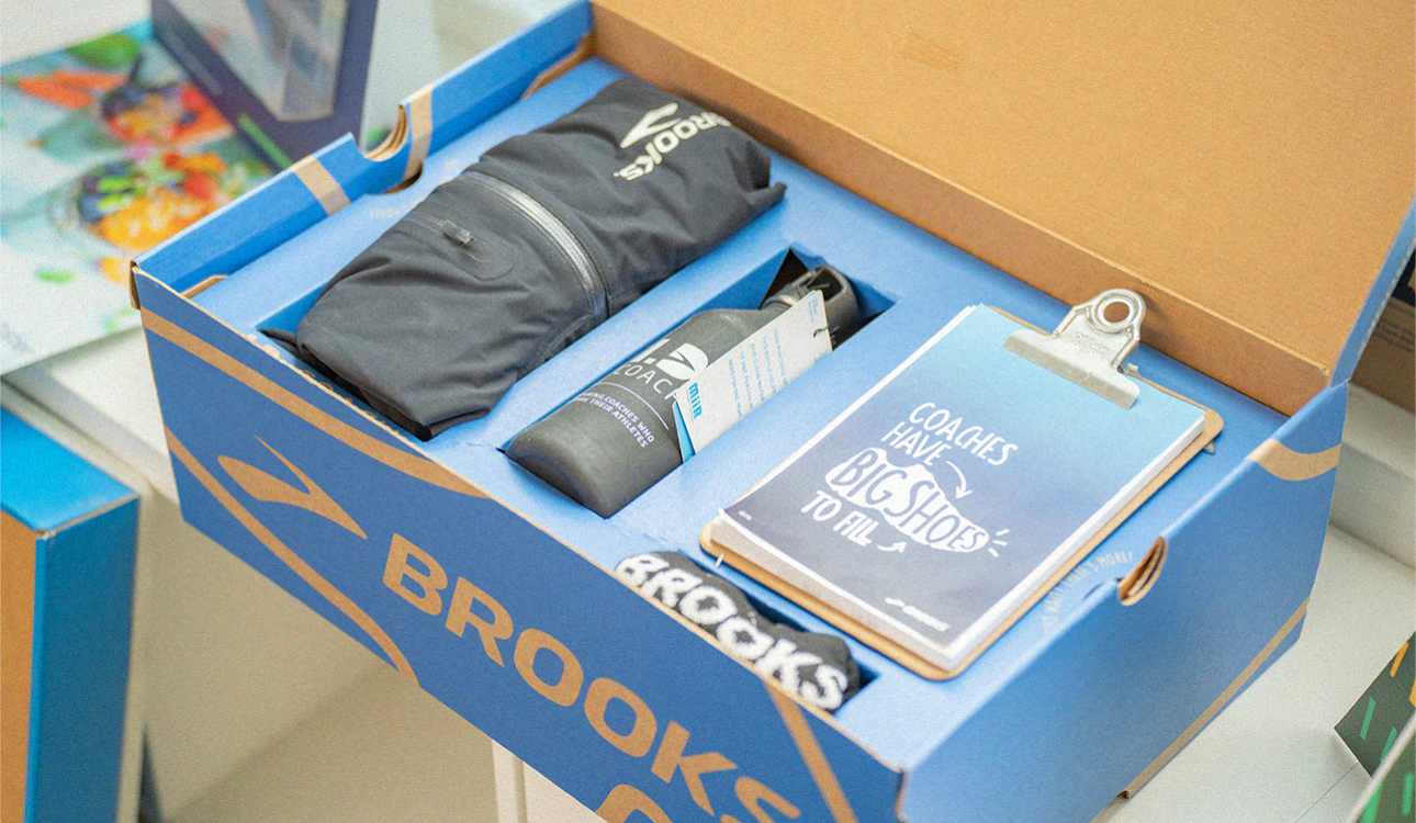 A Brooks Running box features a branded zip-up jacket, water bottle, buff, and coach clipboard nestled into custom-fit places.