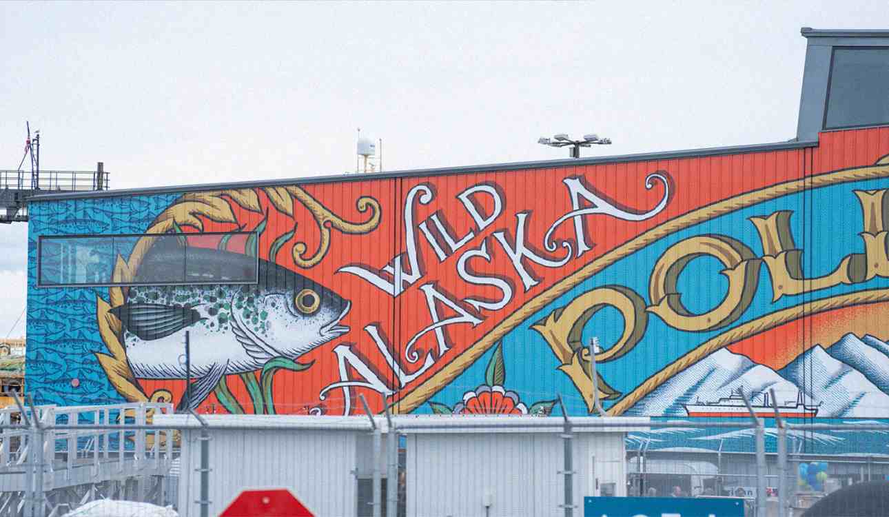 A colorful mural advertising wild Alaska pollock covers an entire corrugated-metal wall on a seaside building.